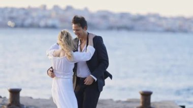 Bir adam ve bir kadın iskelede suyun kenarında duruyorlar. Adam takım elbise giyiyor ve kadın beyaz bir elbise giyiyor. Birbirlerine eşlik etmekten zevk alıyor gibiler.