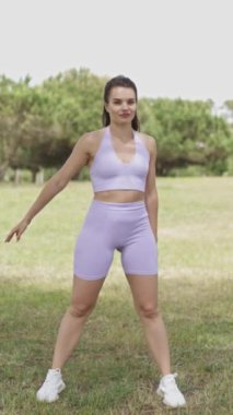 Mor bluzlu ve şortlu bir kadın çimen tarlasında dans ediyor. Gülümsüyor ve eğleniyor.