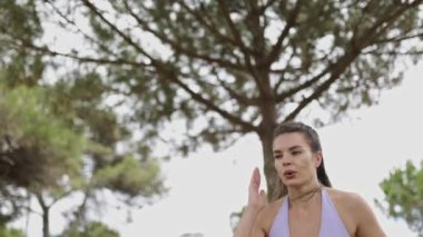 Arka planda ağaçlar olan bir kadın parkta koşuyor. Mor bir bluz giyiyor ve eli yüzünde.