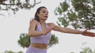 Bir kadın parkta yoga yapıyor. Mor bir bluz giyiyor ve gökyüzüne bakıyor.