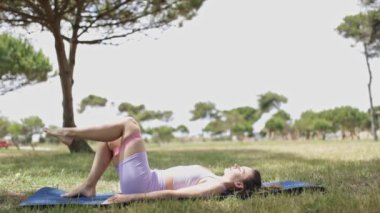 Bir kadın parkta mindere yoga pozu veriyor. Mor bir bluz ve pembe şort giyiyor.