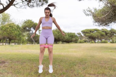 Mor spor kıyafetli bir kadın parkta dikilirken bir direniş bandıyla bacak egzersizi yapıyor..