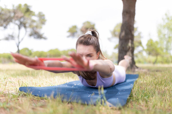 A young woman lies on a blue mat in a park, stretching her arms with a red resistance band.