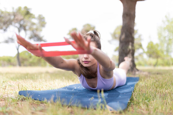 A young woman lies on a blue mat in a park, stretching her arms with a red resistance band.