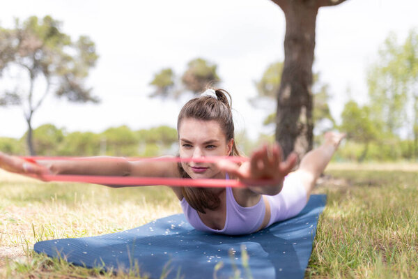 A young woman lies on a blue yoga mat in a grassy park, stretching her arms with a pink resistance band.
