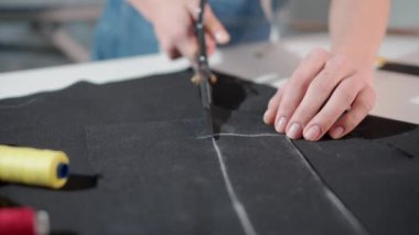 Bir kadın makasla kumaş kesiyor. Makas, bir makara ipliğin yanındaki masada.