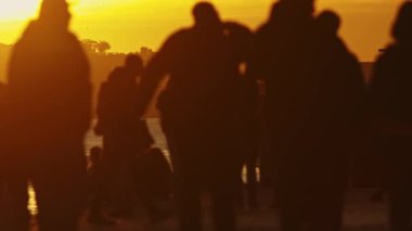 Gün batımında bir grup insan sahilde yürüyor. Halkın siluetleri turuncu gökyüzüne karşı serin ve huzurlu bir atmosfer yaratıyor. Kumsal boş.