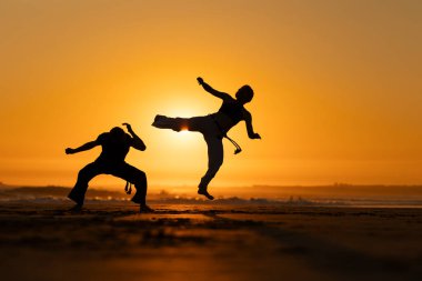 Gün batımında iki kişi sahilde karate çalışıyor. Bir tanesi havada tekme atıyor.