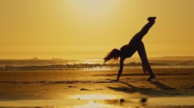 Bir kadın gün batımında sahilde capoeira yapıyor. Sahne sakin ve sakin, arka planda güneş batıyor ve ön planda okyanus dalgaları.