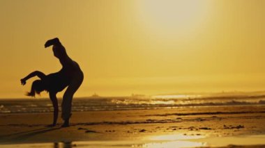 Bir kadın gün batımında plajda tai chi yapıyor. Sahne sakin ve sakin. Arka planda güneş batıyor ve dalgaların sesi ön planda.