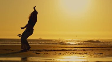 Sahilde bir kadın duruyor, arka planda güneş batıyor. Siyah gömlek ve beyaz pantolon giyiyor.