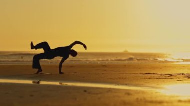 Bir adam kumsalda dövüş sanatları çalışıyor. Gökyüzü turuncu ve su sakin.