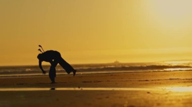 Kapoeira - adam kumsalda dövüş sanatları çalışıyor. Gökyüzü turuncu ve su sakin.