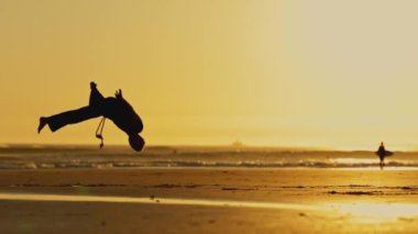Gün batımında bir adam kumsalda dövüş sanatları çalışıyor. Sahne huzurlu ve sakin, erkek hareketleri sakin ve odaklanmış hissi yaratıyor.