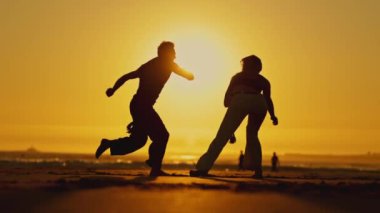 Gün batımında iki kişi kumsalda dövüş sanatları çalışıyor. Güneş arka planda batıyor, sahne üzerine sıcak bir parıltı saçıyor.