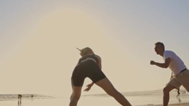 Sahilde gün batımında iki kişi capoeira yapacak. Kadın eğiliyor ve adam amuda kalkıyor. Kumsal boş ve gökyüzü temiz.