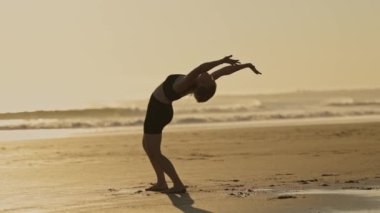 Bir kadın sahilde akrobatik egzersizler yapıyor. Okyanus kenarında gün batımı.