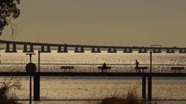Köprü, üzerinde birkaç kişinin yürüdüğü bir su kütlesini kapsar. Arka planda güneş batarken sahne huzurlu ve huzurlu.