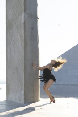 Genç kadın şehir ortamında dans ediyor, siyah bir elbise giyiyor ve dans ayakkabıları giyiyor.