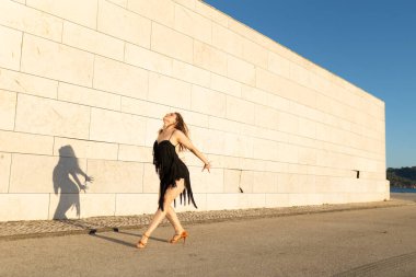 Genç bir kadın sokakta beyaz bir duvara karşı Latin dansı yapıyor.