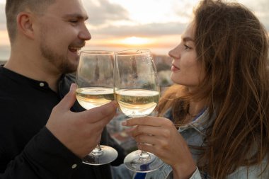 Bir erkek ve bir kadın gün batımında çatıda romantik bir anın tadını çıkarıyorlar, beyaz şarap bardaklarını tokuşturuyorlar ve gülümsüyorlar.