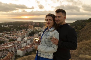 Çiftler, bir şehir manzarasında gün batımını izlerken, birbirlerine sarılıp şarap içiyorlar.