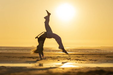 Genç bir kadın gün batımında sahilde Capoeira hareketi yapıyor. Güzel bir siluet yaratıyor.