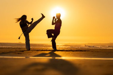 Sahilde batan güneşin gölgesinde Capoeira hareketleri üzerinde çalışan iki kişi.