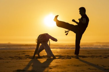 Gün batımında sahilde capoeira yapan bir kadın ve erkeğin siluetleri.