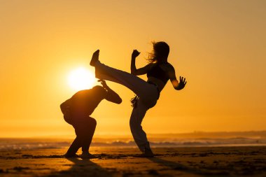 Gün batımında sahilde capoeira yapan bir adamla bir kadının siluetleri.