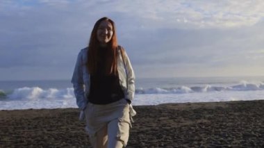 Gülümseyen kadın resimli bir plajda yürüyor. Arka planda yavaşça dalgalar çarpıyor.