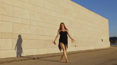 Siyah elbiseli bir kadın beyaz bir duvara karşı gösteri yapıyor.