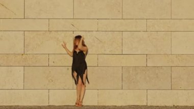 Siyah elbiseli dansçı bej tuğla bir duvara karşı koreografi yapıyor.