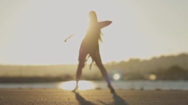 Kadın, arka planda batan güneş olan bir su kütlesinin yakınındaki asfalt bir yüzey üzerinde dans rutini uyguluyor.