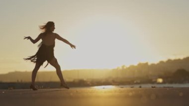 Dans kostümü giymiş bir kadın beton bir yüzeyde arka planda batan güneş olan bir su kütlesinin yanında salsa yapıyor.