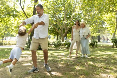 Baba, karısı ve annesi onları izlerken oğlunu parkta döndürüyor.