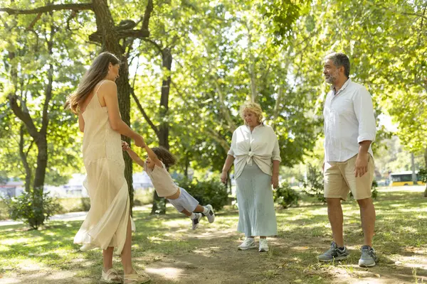 stock image Cheerful multigenerational family enjoying a sunny day in the park, with a mother swinging her child