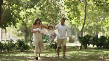 Genç bir aile, yeni doğmuş bir bebeği taşıyan bir anne, ve küçük çocuklarının elini tutan bir baba, güneşli bir parkta sakin bir gezintinin tadını çıkarıyor.
