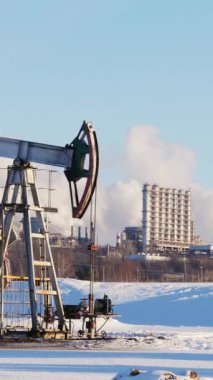 Petrol pompası, kar kaplı bir petrol sahası ve rafineri zeminiyle kış manzarasında çalışıyor.