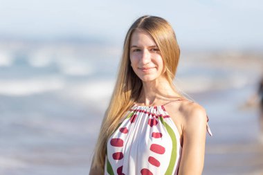 Sarışın kadın sahilde gülümsüyor. Arka planda okyanus dalgaları var.