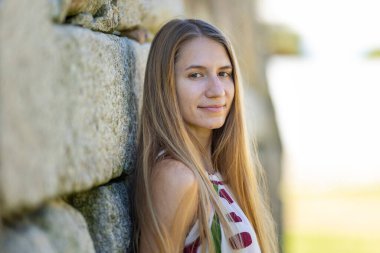 Genç sarışın kadın güneşli bir günde taş bir duvara yaslanıyor.