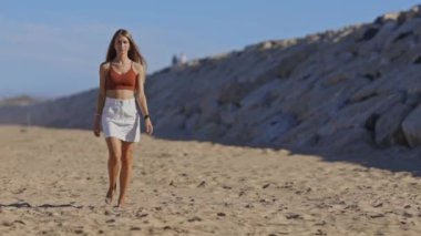 Genç bir kadın güneşli bir sahilde kameraya doğru yürüyor.