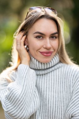 Kafasında güneş gözlüğü olan, beyaz balıkçı yaka kazak giyen, gülümseyen ve saçlarına dokunan çekici genç bir kadın.