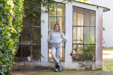 Genç bir kadın, yaşlı bir seranın önünde duruyor. Duvara yaslanmış, kolları arkada.