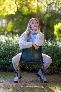 Genç bir kadın parkta bir sandalyede oturuyor, beyaz bir kazak ve yırtık kot pantolon giyiyor.