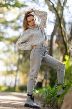 Genç bir kadın parkta poz veriyor, saçlarına dokunuyor, kot pantolon ve süveter giyiyor.