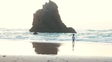 Küçük çocuk kumlu bir sahilde okyanus dalgalarına doğru koşar. Islak kumda yansıyan büyük bir kaya oluşumu.