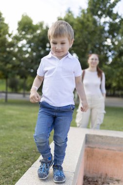 Beyaz gömlekli ve kot pantolonlu genç bir çocuk beton duvarda yürüyor. Arkasında bir kadın var.