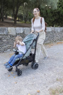Bir kadın içinde çocuk olan bir bebek arabasını itiyor. Çocuk gülümsüyor ve kadın da gülümsüyor. Sahne huzurlu ve mutlu.