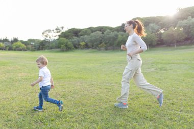 Anne ve oğul parkta çimenlerde koşuyorlar, güzel bir yaz gününün tadını çıkarıyorlar.